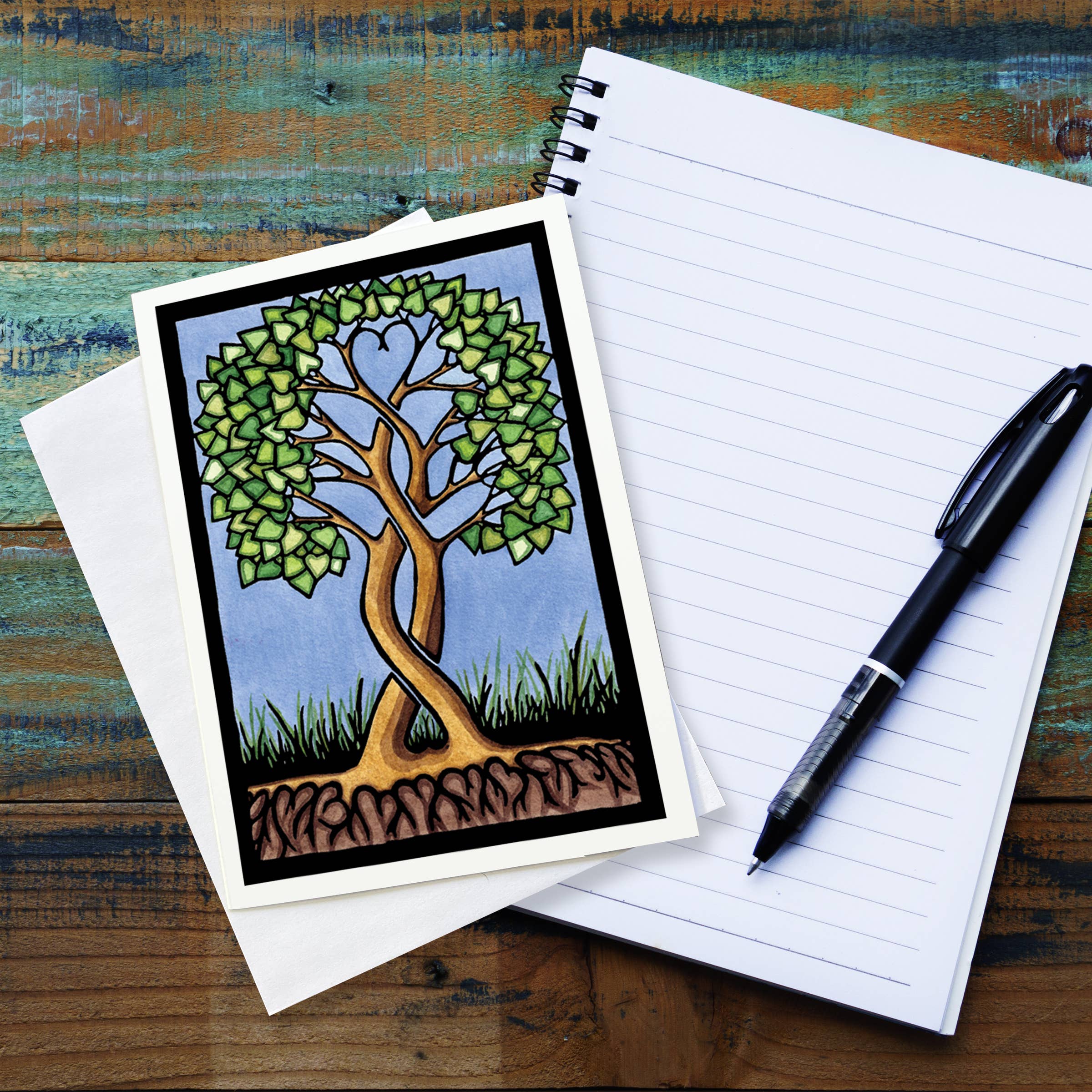 A Card: Together Forever Tree by Sarah Angst 5x7 rests on a blank envelope next to an open lined notebook and a black pen on a wooden surface. Made in America.