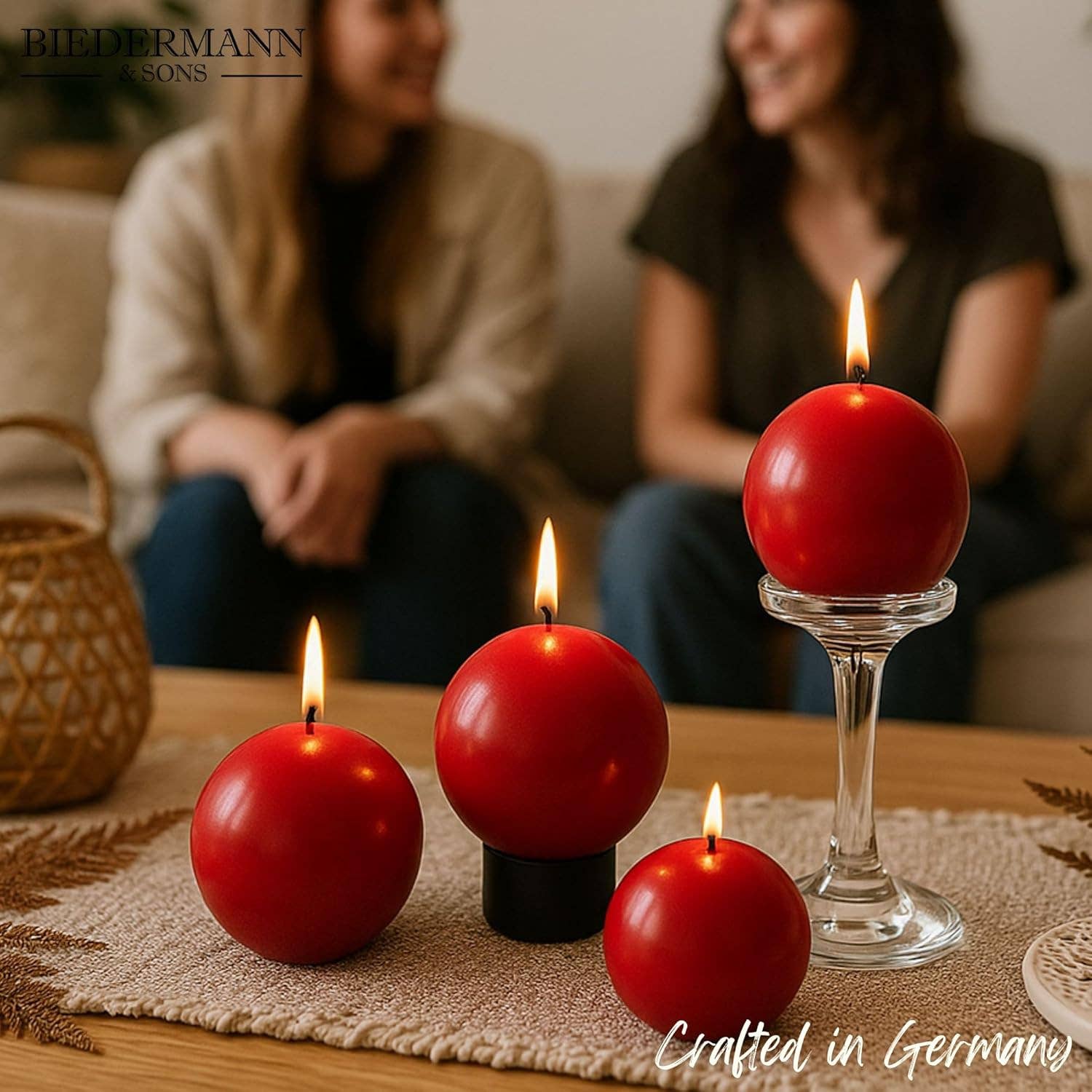Four Cranberry Red Ball Candles, 2.5" diameter, glow brightly in the foreground—three on a table and one on a glass stand—while two people chat on a couch in the background, setting a cozy scene with decorative candlelight.