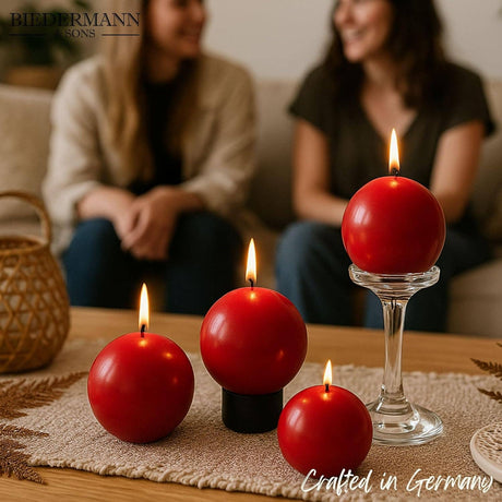 Four Cranberry Red Ball Candles, 2.5" diameter, glow brightly in the foreground—three on a table and one on a glass stand—while two people chat on a couch in the background, setting a cozy scene with decorative candlelight.