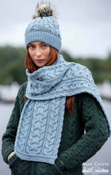 A woman stands outdoors in a dark green textured sweater, wearing the Chunky Cable Hat - Mermaid (One Size), made in Ireland from merino wool, paired with a matching scarf.