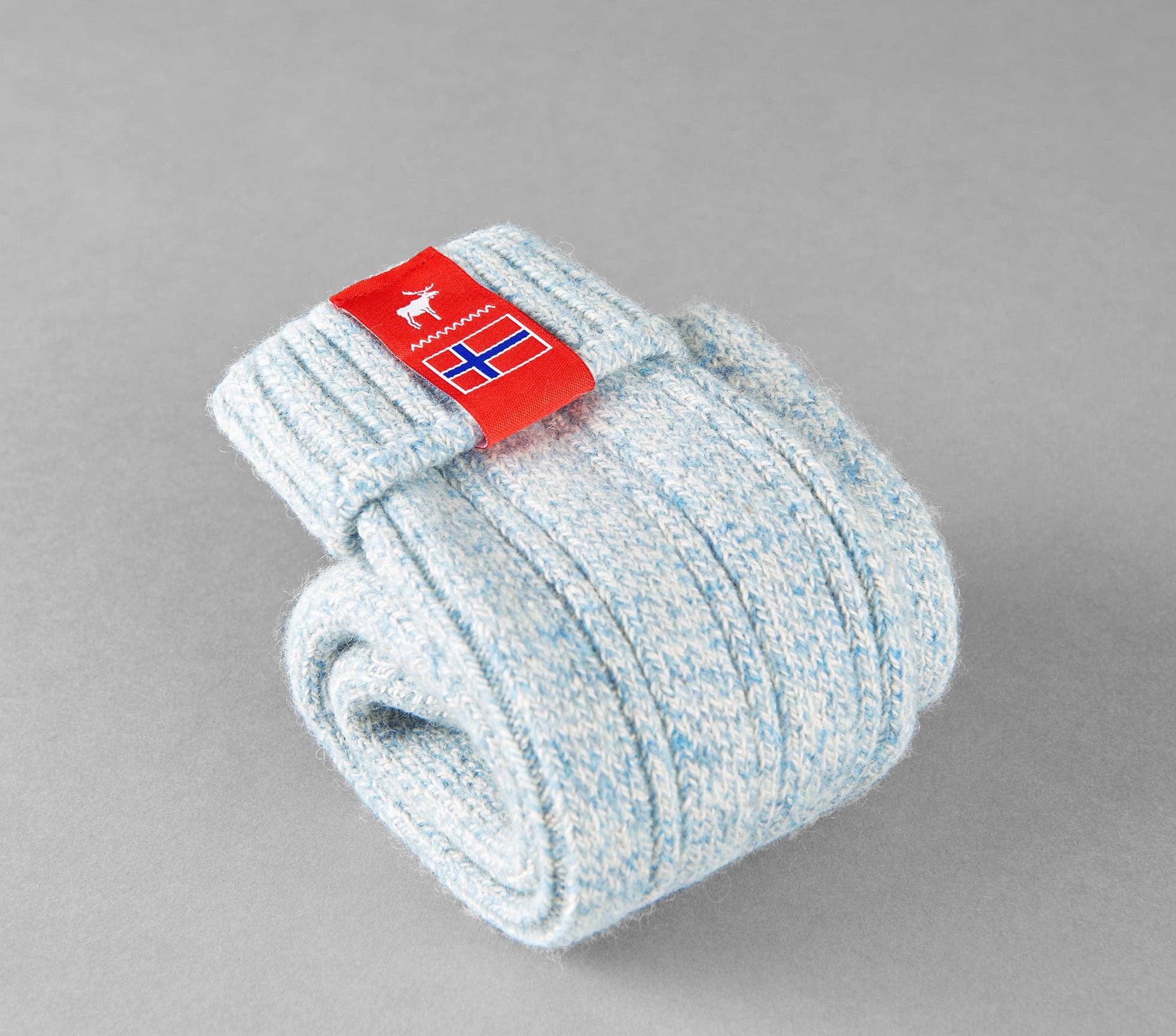 Discover the Norwegian Fjord Socks, a cozy light blue knit pair ideal for Nordic pampering, adorned with a red tag showcasing a deer and a flag icon set against a gray background.