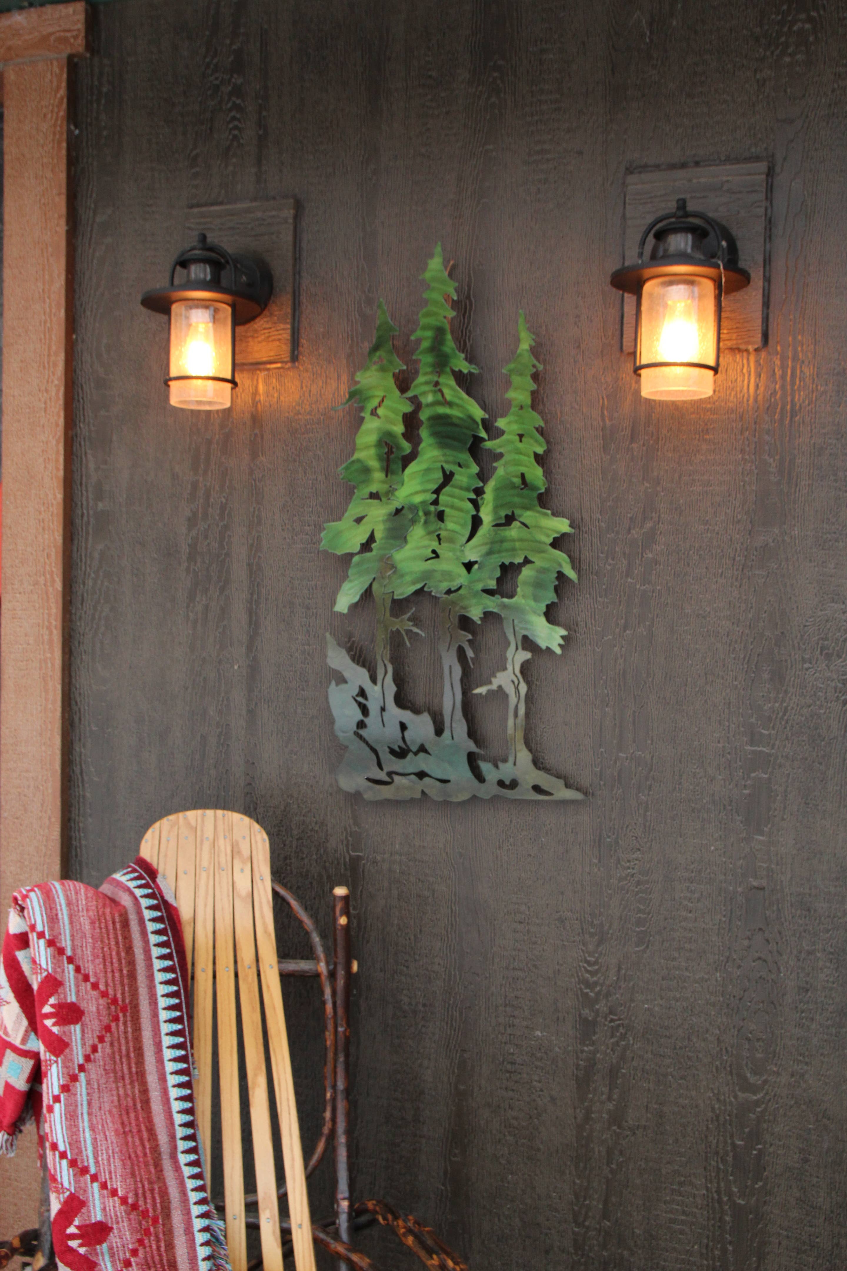 A sled and a patterned blanket rest against a wall adorned with 22" Pine Tree Metal Wall Art, flanked by two rustic lantern lights.