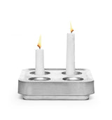 Two white candles of different heights stand in the Stumpastaken Fyran Four Candle Holder – Square, made from 100% recycled material with a white powder-coated finish, highlighting sustainable decor against a plain white background.