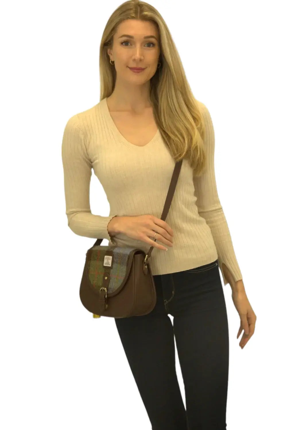 A woman with long blonde hair wears a cream ribbed sweater and dark pants, carrying a Harris Tweed Saddle Bag in dark brown with a blue, brown, and yellow tartan wool flap.
