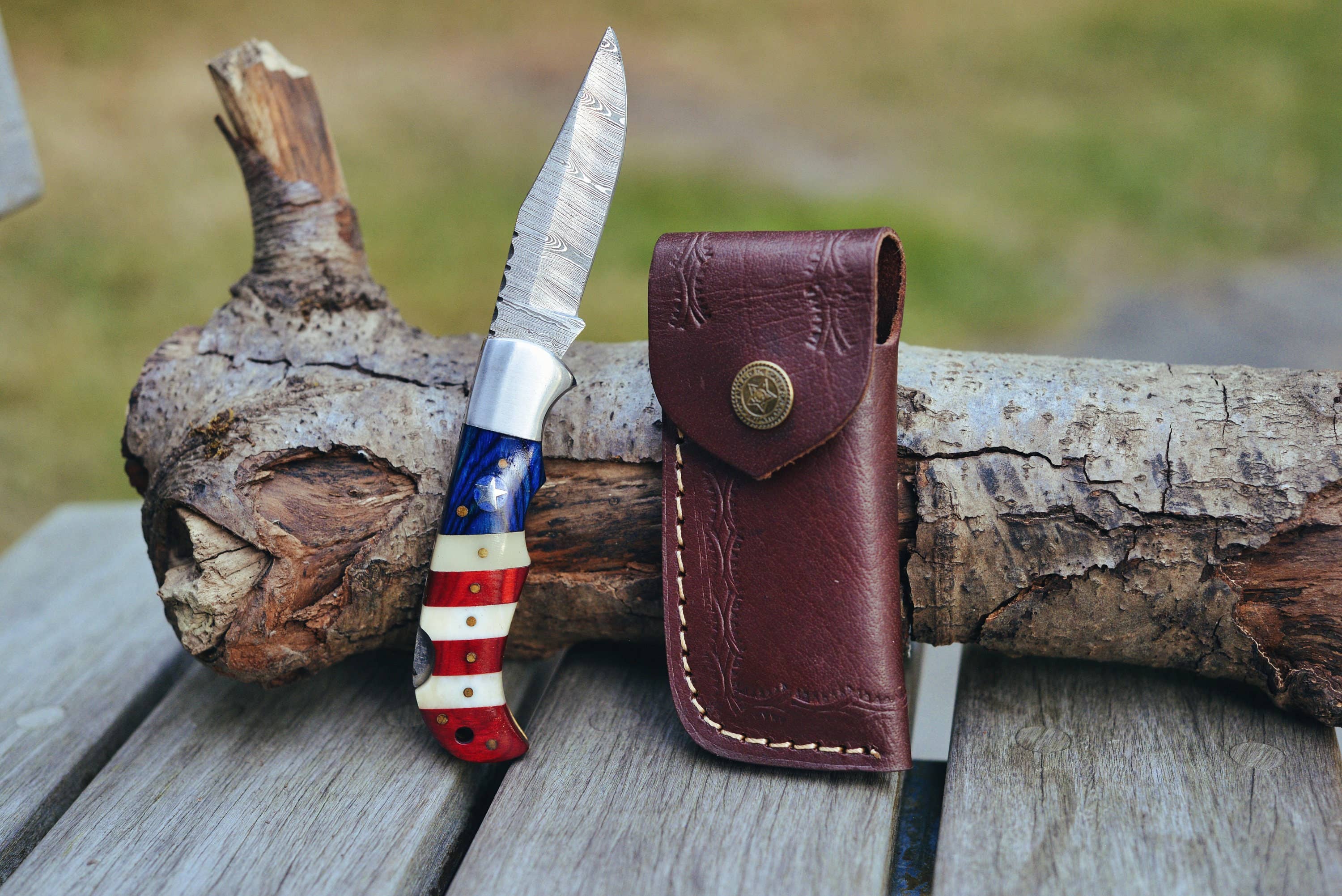 A USA Flag Damascus folding pocket knife with a red, white, and blue handle is placed against a log on a wooden surface. A brown leather sheath, perfect for personalized gifts, is positioned vertically beside it.