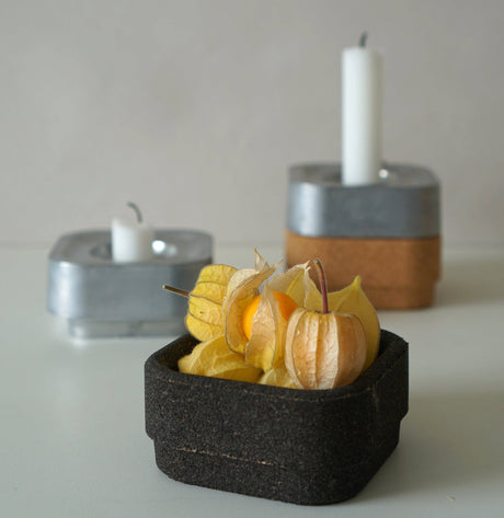 A square black container with yellow physalis fruit is in the foreground; behind it are two Stumpastaken Ettan candle holders (white, 100% recycled, 8.8x8.8x4 cm) with white candles—one lit and one unused—highlighting sustainable design.