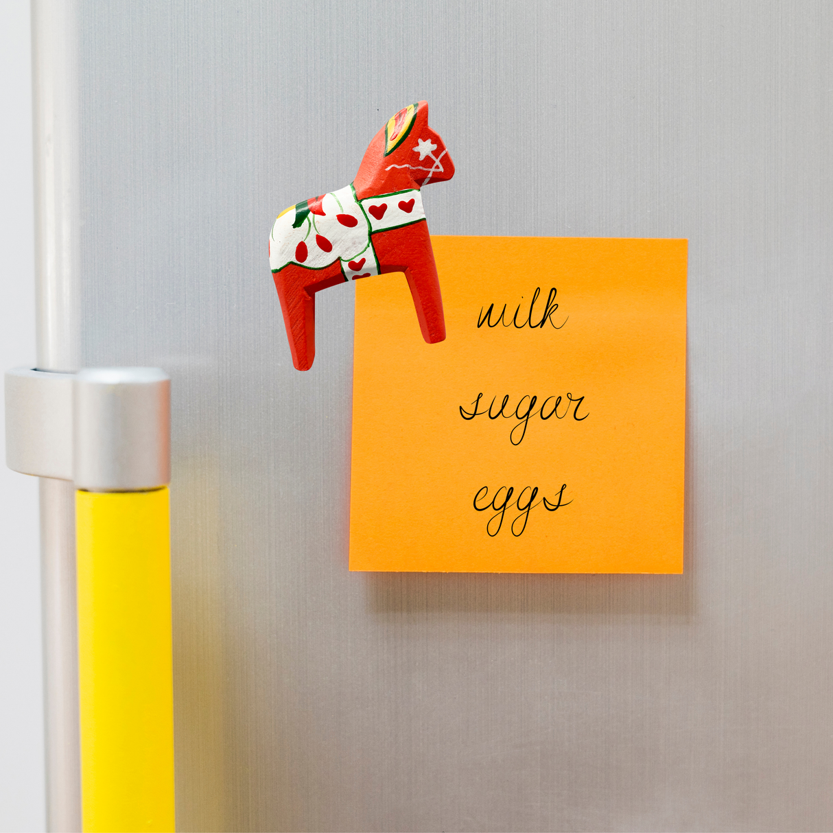 An orange sticky note with milk, sugar, and eggs written on it is attached to the fridge with a European Heritage Gifts Magnet: Red Swedish Dala Horse Refrigerator Magnet (3" tall by 2.5" long), adding a charming Scandinavian souvenir touch.