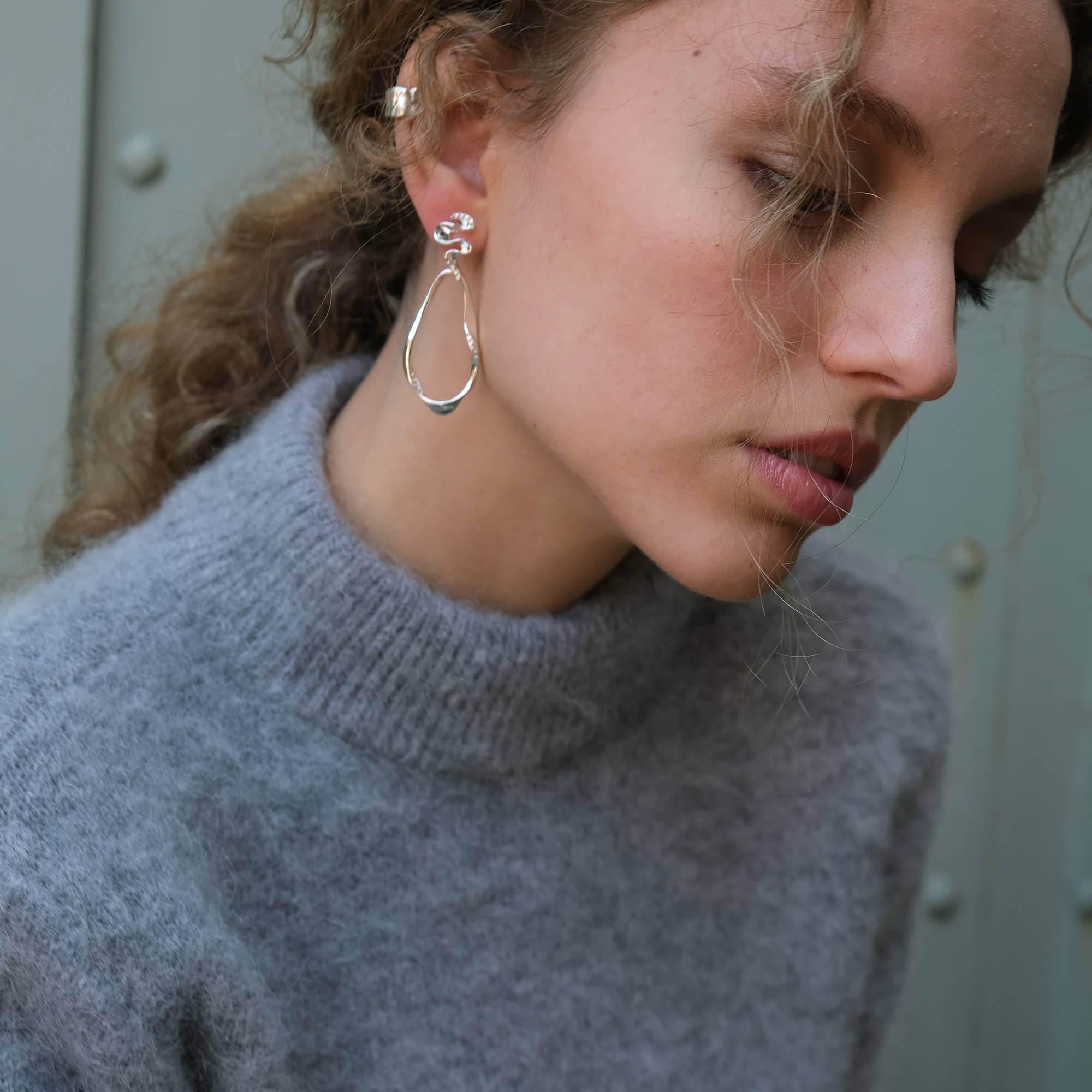 Woman with curly hair wears a grey sweater and Blowing Ribbon Silver Dangle Earrings by A&C Oslo, shown in a close-up side profile against a neutral background.