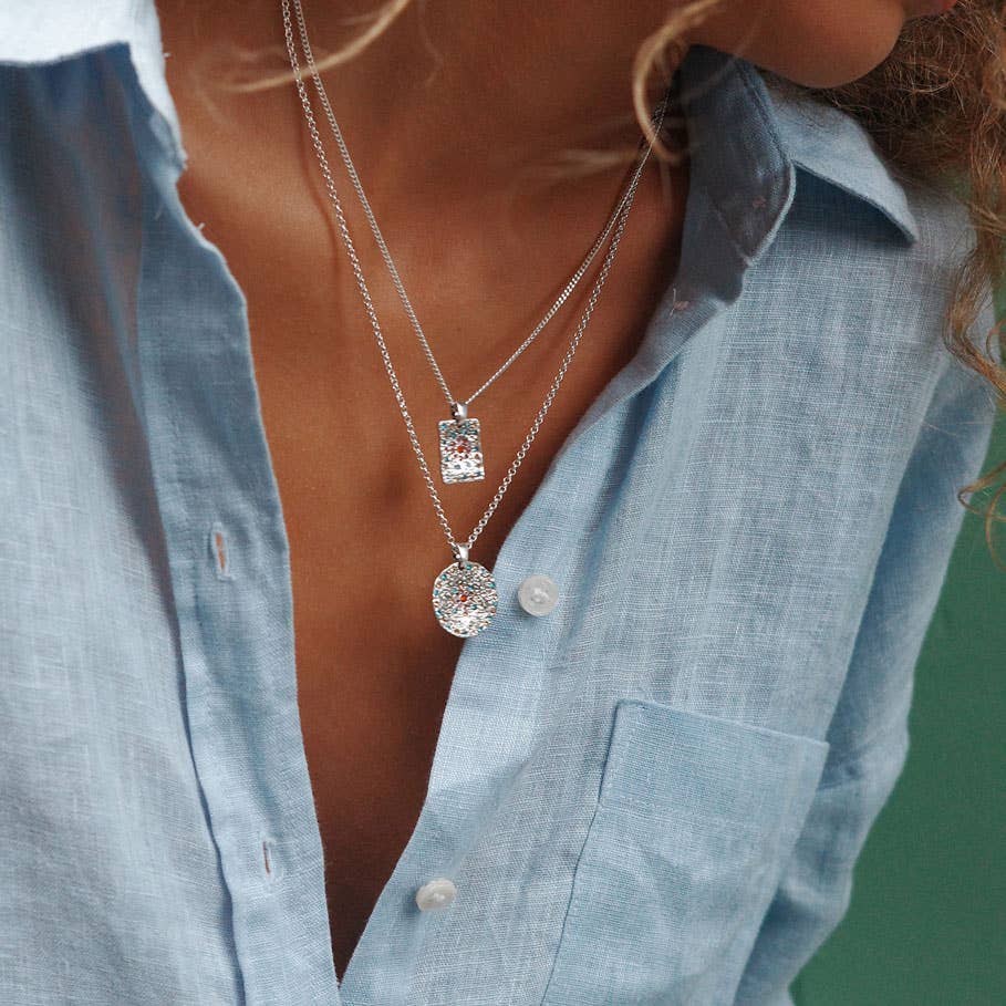 Accessorizing their light blue button-up shirt, a person wears two silver necklaces: one with vibrant colorful stones and the A&C Oslo Vibrance Silver Coin Necklace, adding a touch of elegance.