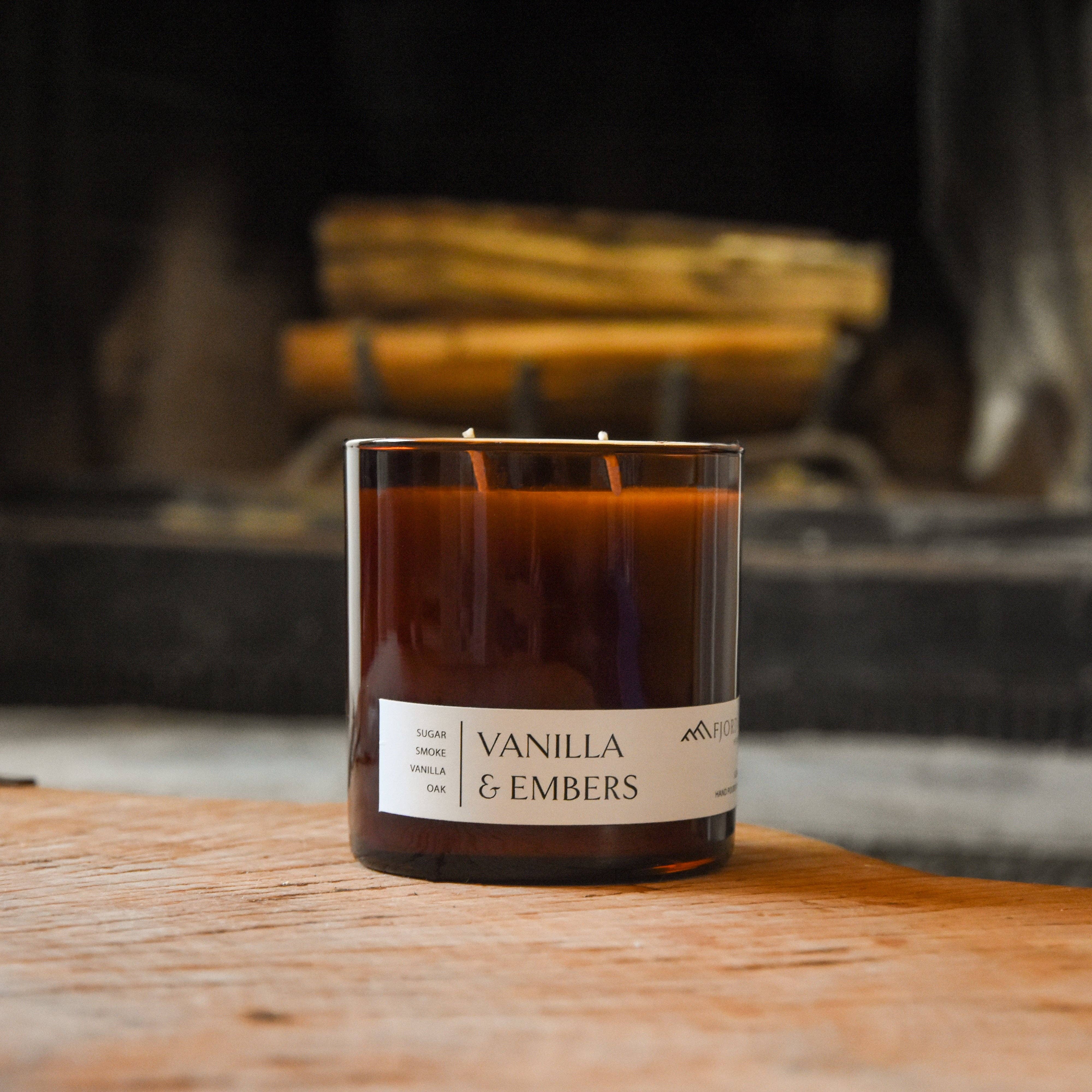 A brown glass 4oz Vanilla Charcoal Oud soy candle labeled "Candles: Vanilla & Embers Premium Soy Candle" with two wicks sits elegantly on a wooden surface, against the rustic backdrop of stacked logs.