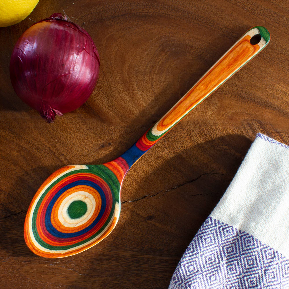 A birch serving spoon with colorful concentric rings is on a wooden surface beside a red onion, a lemon, and a folded white towel with a purple geometric pattern.