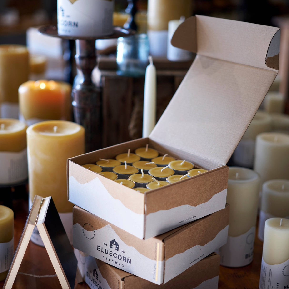 An open box of Pure Beeswax Tea Light Candles in raw or clear cups sits on stacked boxes, surrounded by various other candles such as elegant pillar candles, on a wooden surface.