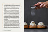 A hand sifts powdered sugar over three cream-filled Danish pastries on a dark surface. To the left lies "Book: Scandinavian from Scratch" by Penguin Random House, open to a page about the pastries' history in Scandinavian baking.