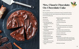A chocolate cake with one slice missing sits on a dark surface. The *Cozy Christmas Cookbook 50 Recipes to Rediscover the Magic of the Season* by Taylor Vance features Mrs. Clauss Chocolate On-Chocolate Cake, offering a modern twist to holiday recipes. Nearby are a cake server and ingredients.