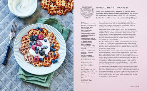 A plate of heart-shaped waffles adorned with yogurt and berries sits on a gray tablecloth next to a green napkin, capturing the essence of Scandinavian cuisine. Adjacent, a bowl with creamy goodness enhances this Nordic treat, while the ScandiKitchen Cookbook entices curiosity on the right.