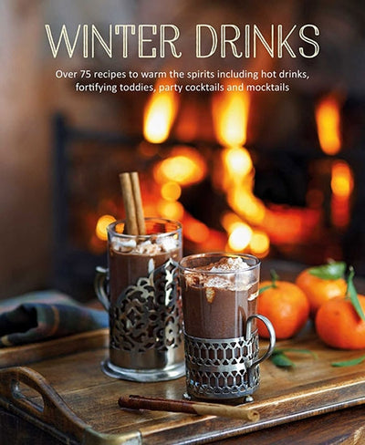 Two glasses of hot chocolate with cinnamon sticks rest on a tray, placed before a crackling fireplace, accompanied by oranges. Text reads: "Book: Winter Drinks - Over 75 recipes to warm the spirits, featuring mulled wine delights.