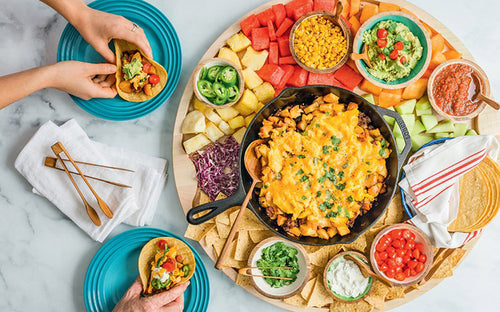 An array of taco ingredients is beautifully displayed on a platter, ideal for family meals, featuring cheese, vegetables, salsa, and guacamole. Two hands are putting together tacos on blue plates from the "Big Boards for Families" book.