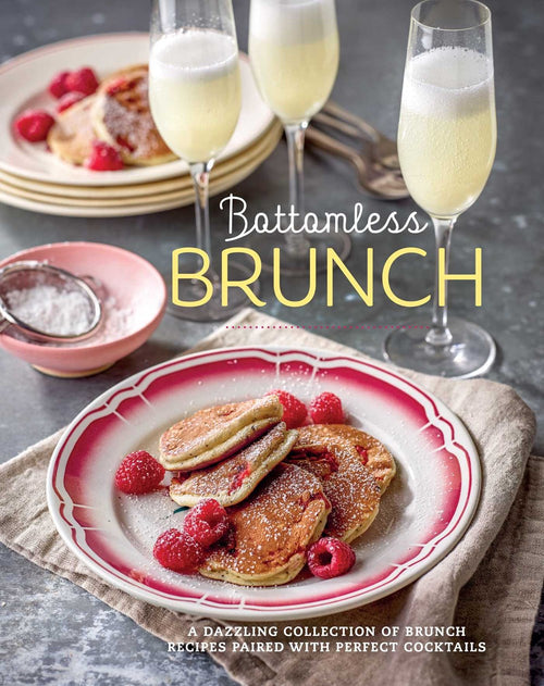 A plate of pancakes with raspberries is set next to boozy cocktails and glasses of champagne on a table, with the elegant display of the book "Bottomless Brunch.