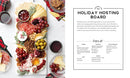 A holiday entertaining board curated with a variety of cheeses, meats, olives, grapes, and spreads features a white card detailing the menu on the right. It's ideal for highlighting seasonal recipes as featured in "Around the Board.