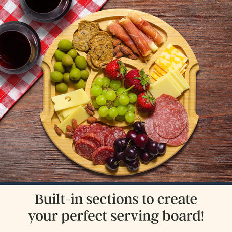 Round bamboo cutting board with compartments containing crackers, cheese, grapes, strawberries, olives, nuts, and charcuterie, displayed beside two glasses of wine.