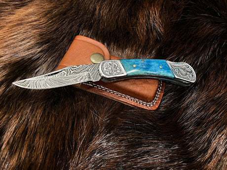 The Knife: Dyed Blue Bone 6.5 Damascus Steel features an engraved Damascus blade and a blue handle, displayed on a brown leather sheath atop fur.