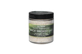 A clear jar from the SALTVERK Gift Set features hand-harvested Icelandic Birch Smoked Sea Salt, with a green label and black lid. The set includes Flaky Sea Salt, Lava Salt, Birch Smoked Salt, and Arctic Thyme Salt from the Westfjords.