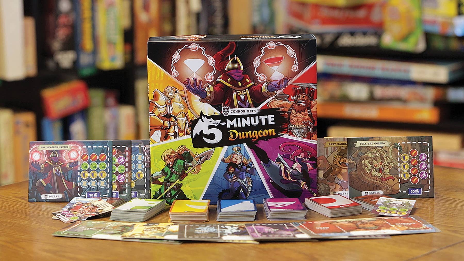 The cooperative card game, Game: 5 Minute Dungeon Board Game, features colorful cards and character mats arranged on a wooden table, with the game box prominently showcased in the background.