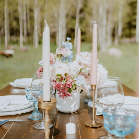 A rustic outdoor table features white plates, a vase of pink and white flowers, and eco-friendly dripless handmade beeswax taper candles in brass holders against a blurred backdrop of trees and grass.