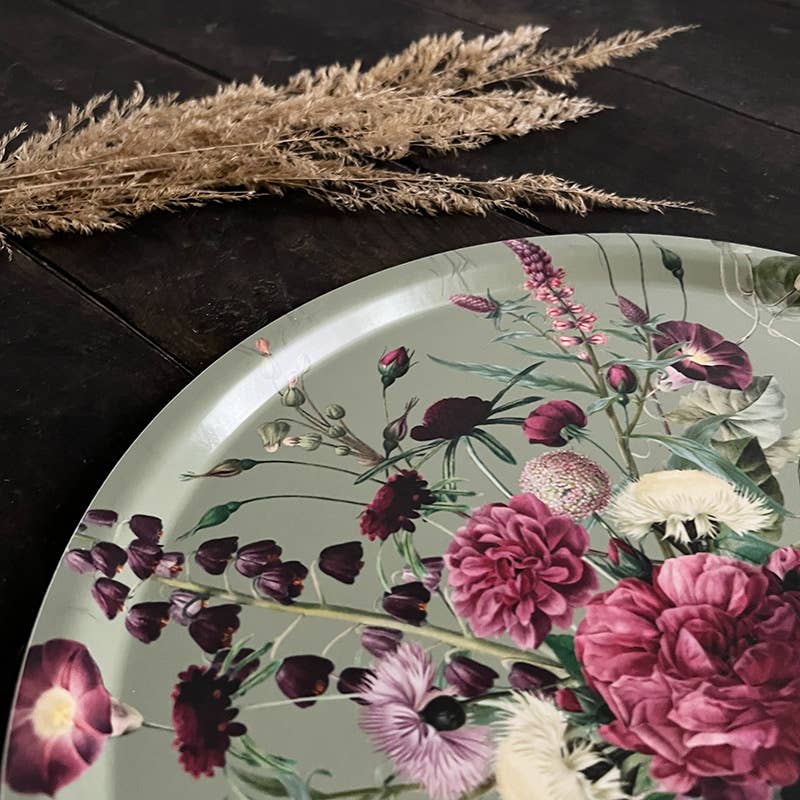 Round serving tray with colorful floral pattern placed on a dark wooden table next to dried grass stems.