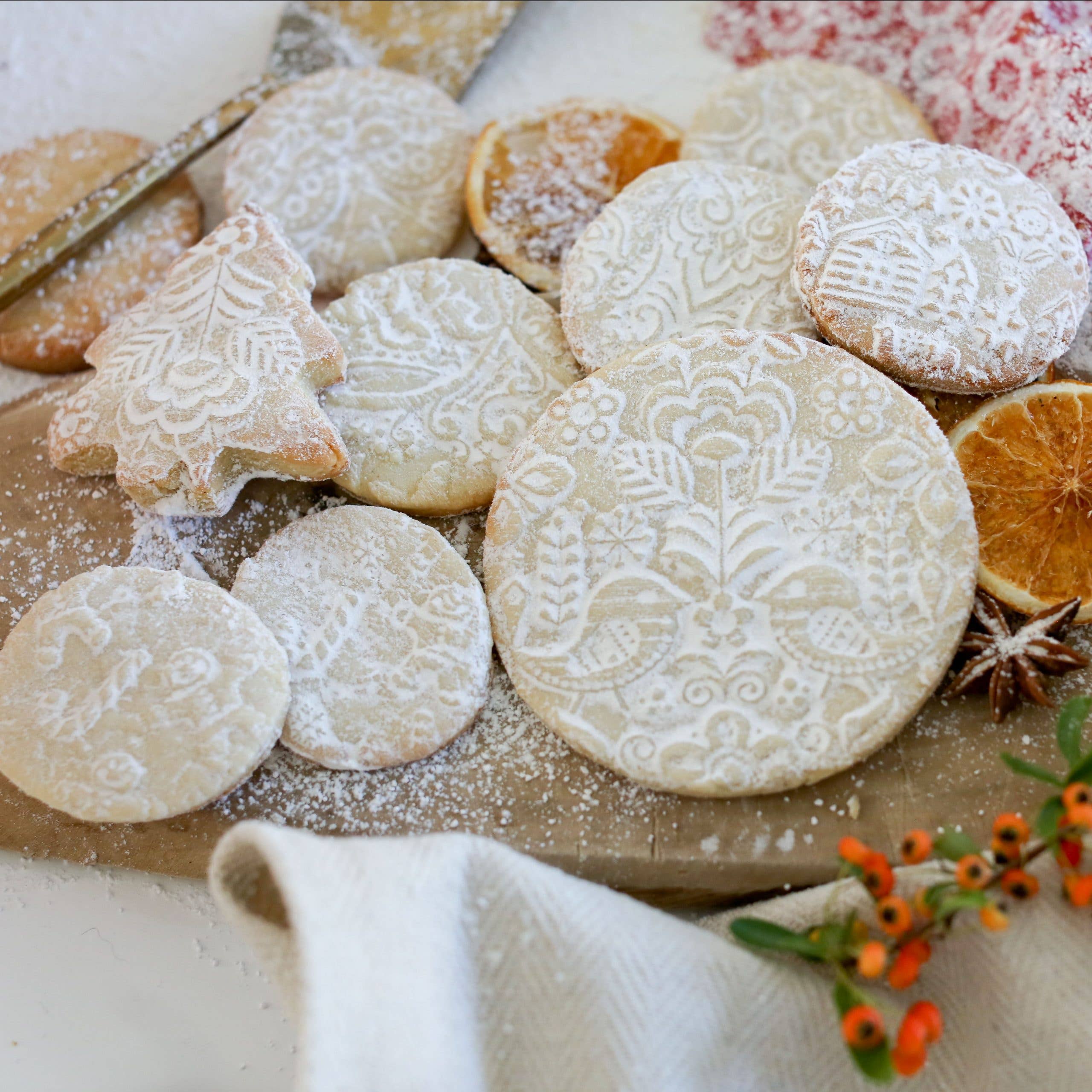 Cookies, made with the Autumn Leaves Embossing Rolling Pin, sit on a wooden board. Theyre sprinkled with powdered sugar and decorated with dried orange slices and berries.