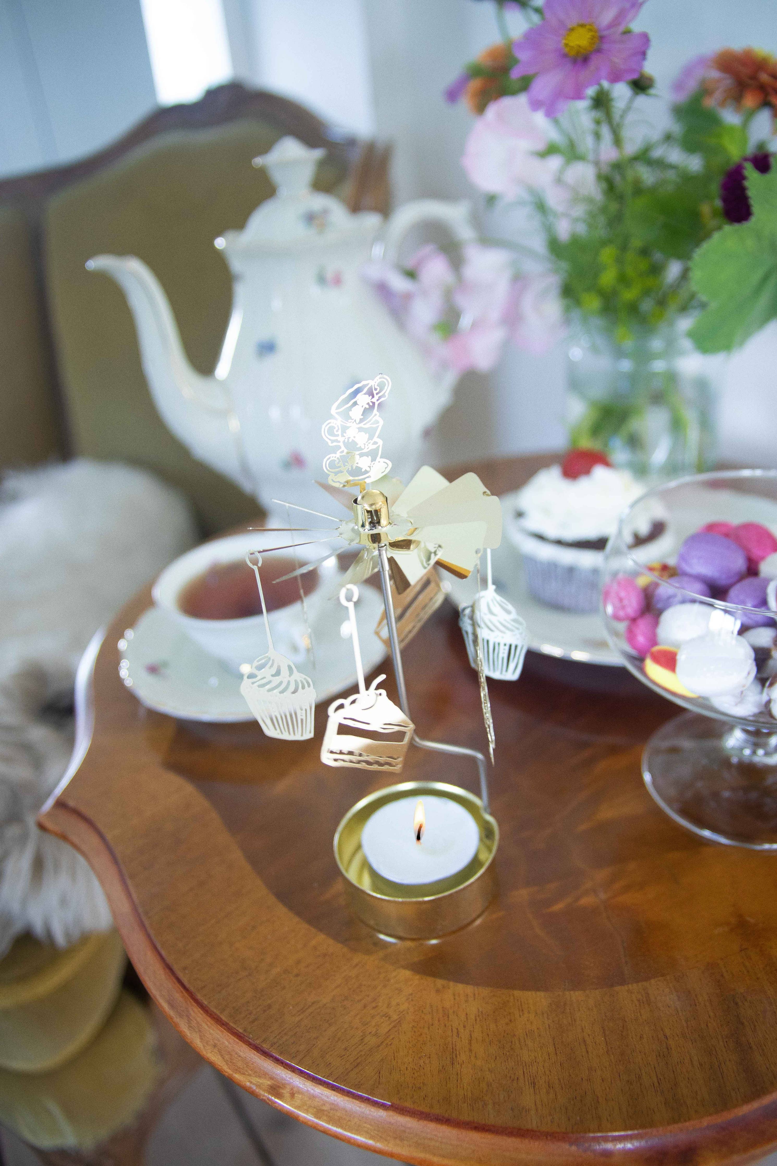 The small table, decorated with various interior details, showcases a silver rotary spinning candle holder named "Candle Holder: Forest Animals," crafted from recycled metal. It is accompanied by a teapot, floral arrangement, cupcake, tea cup, and a bowl of macarons.
