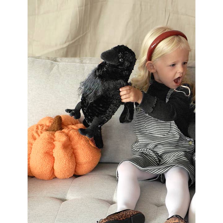 A young child with blond hair, dressed in a checkered dress, white tights, and a red headband, sits on the couch holding the Faire - The Petting Zoo Plush: Raven Wild Onez Raven Stuffed Stands Animal 12″ Tall next to an orange plush pumpkin.
