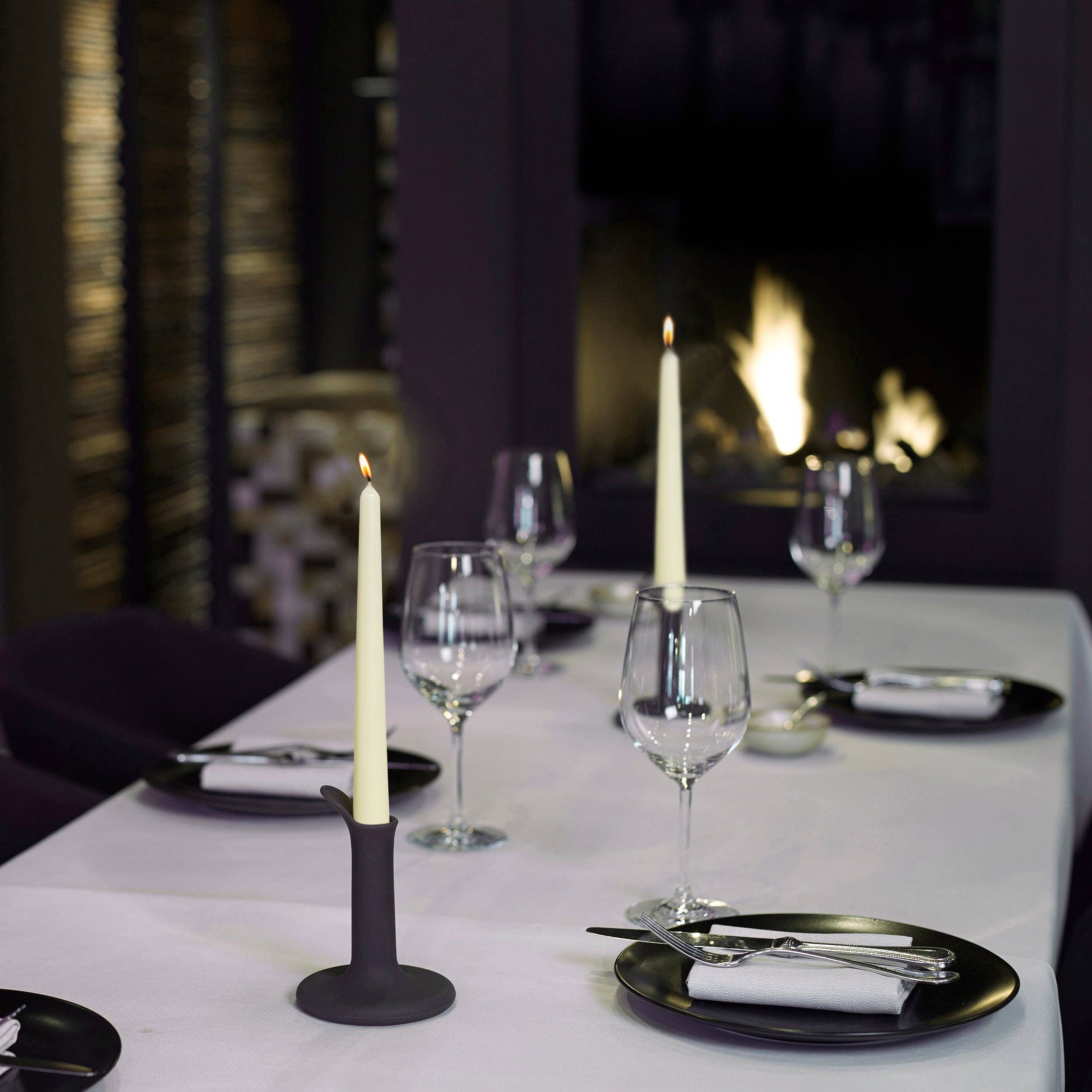 An elegant dining table features a white tablecloth adorned with black plates, silver cutlery, and folded napkins. High-quality white taper candles from the White Tall 10" Taper 4 Pack are used; they are unscented and dripless. These lit candles accompany wine glasses beautifully placed in front of a cozy fireplace.