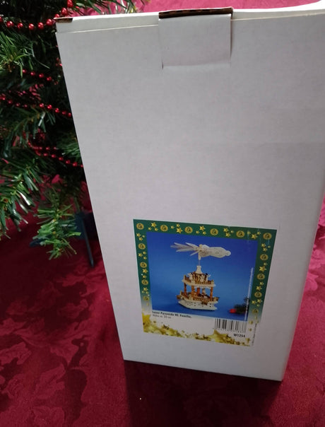 A box featuring the Marketplace24 "Pyramid: Christmas Pyramid Nativity Square with Manger Scene on the Bottom and 4 Angels with Horns on the Top 13'' Tall" is displayed on a red tablecloth next to a decorated Christmas tree, its colorful label depicting a festive German carousel scene.