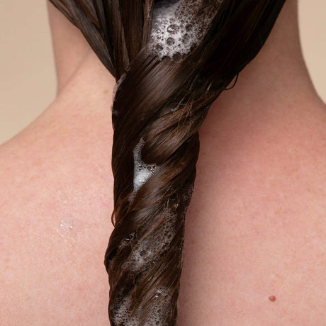 A close-up of the back of a person’s neck shows long, dark, wet hair twisted into a braid and covered in suds from Kitsch Hair: Volumizing Rosemary & Biotin Shampoo 12oz.
