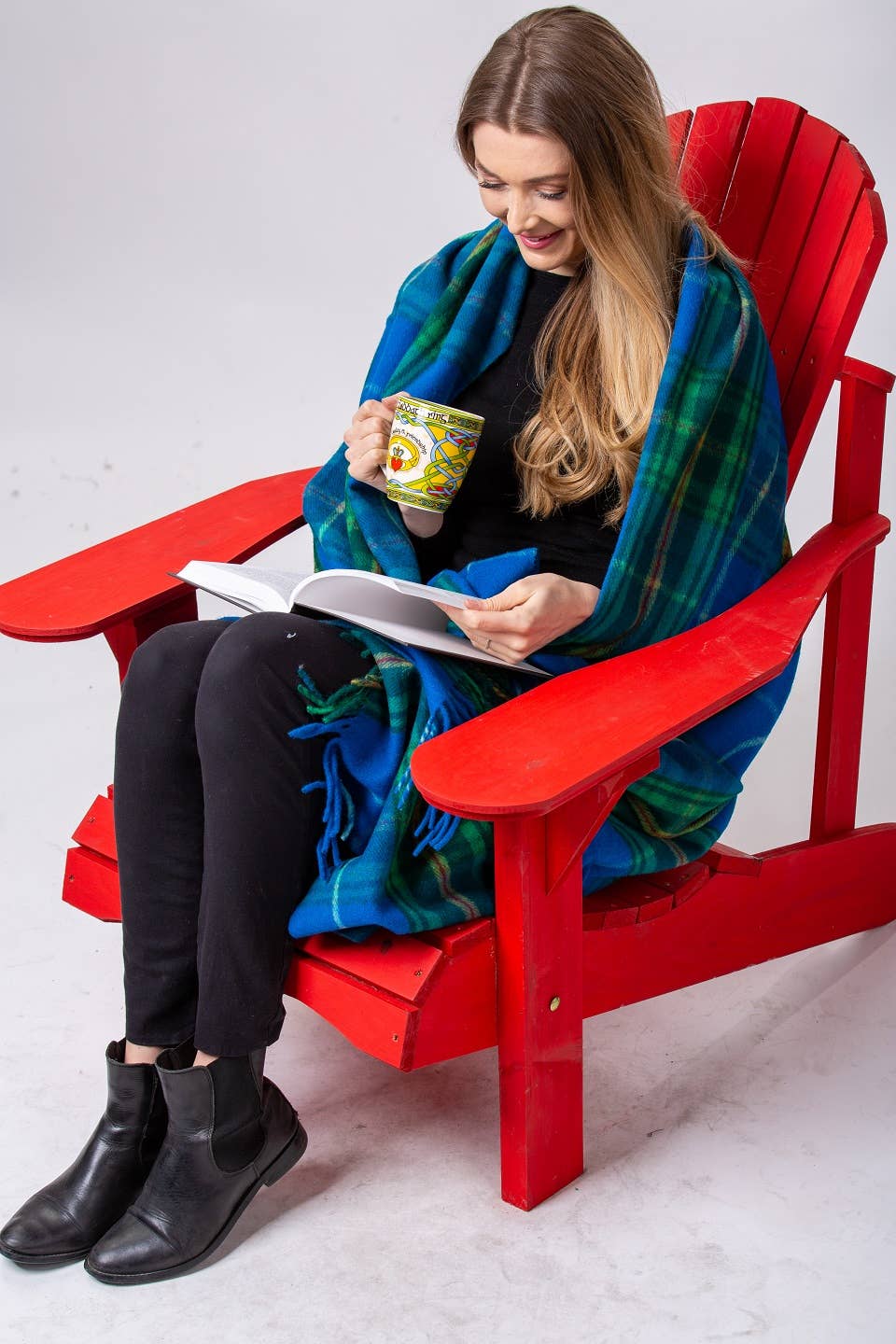 A woman sits on a red chair, wrapped in a Nova Scotia Deluxe Highland Tartan Blanket made of merino wool, holding a mug and reading a book.