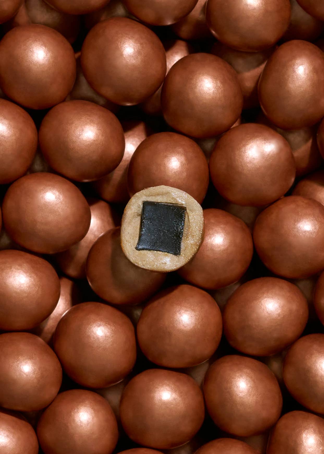 A single tan Candy:CLASSIC CARAMEL Chocolate Coated Licorice: Regular Limited Edition with a black square sits among many round, shiny, chocolate-coated licorice candies.