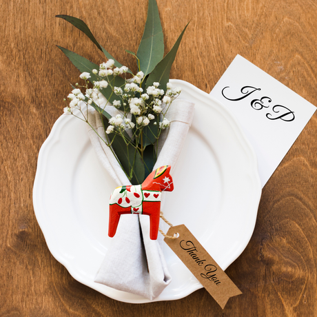 A white plate with a folded napkin, greenery, and flowers is decorated with a European Heritage Gifts Red Swedish Dala Horse Refrigerator Magnet (3" tall by 2.5" long) and a “Thank You” tag. A card inscribed with “J & P” rests on the plate’s edge, bringing a delightful Scandinavian souvenir accent to the table setting.