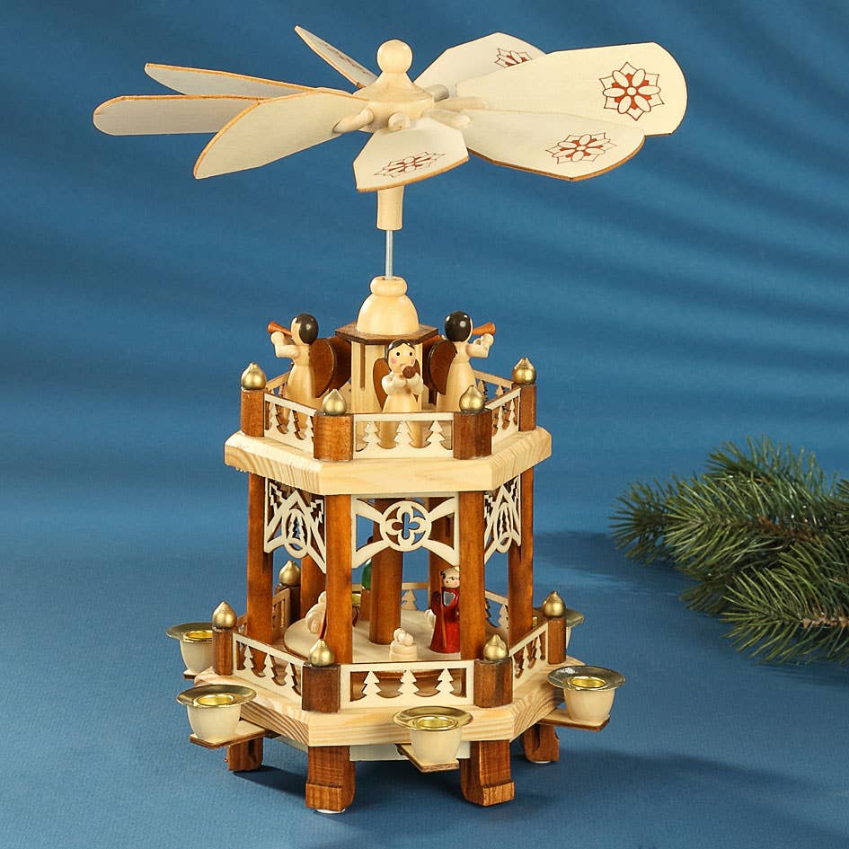 The Pyramid: Nativity Fence Christmas Pyramid, handcrafted in Germany, features carved nativity figures, candle holders, and a rotating fan on top, beautifully displayed with a blue cloth and pine branch.