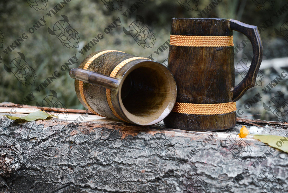 A handcrafted wooden Viking tankard with a vibrant orange finish (16–18 oz) is shown outdoors on a rough tree trunk, one mug upright and the other lying on its side.