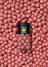 A clear jar of Lakrids by Bülow - Pink Pineapple Danish Chocolate Coated Licorice (125g) sits on a surface covered with the same pink licorice candies.