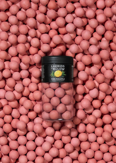 A clear jar of Lakrids by Bülow - Pink Pineapple Danish Chocolate Coated Licorice (125g) sits on a surface covered with the same pink licorice candies.