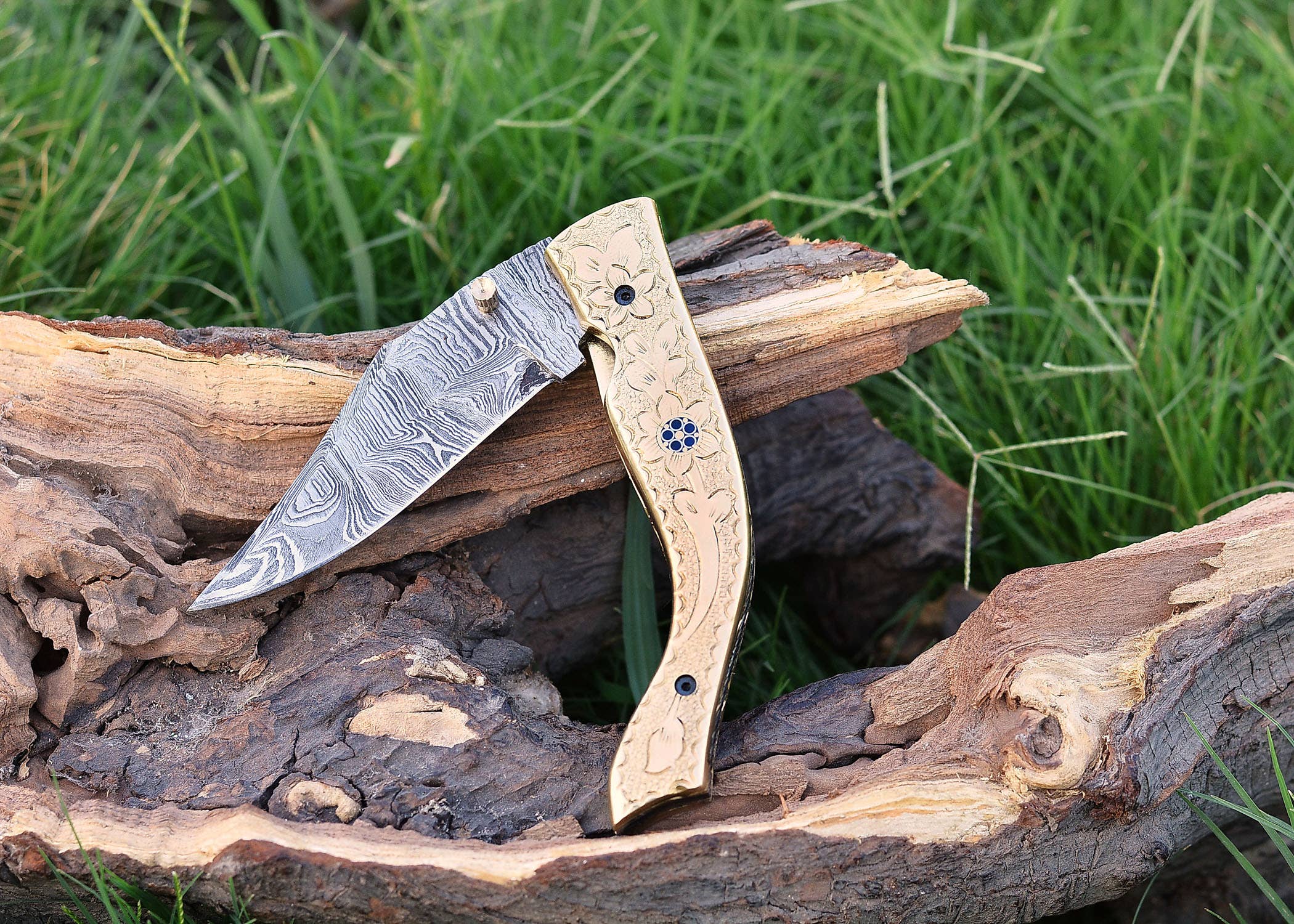 The Exquisite Golden Folding Knife, featuring a patterned stainless steel blade and an intricately carved brass handle, rests on weathered wood against lush green grass.