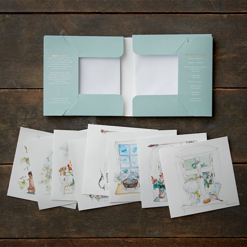 An elegant presentation showcases the "Card Pack: Room with a View," where illustrated postcards of cozy indoor scenes are spread across a wooden surface, complemented by an exquisite square card folder and eucalyptus envelopes adding to the charming display.