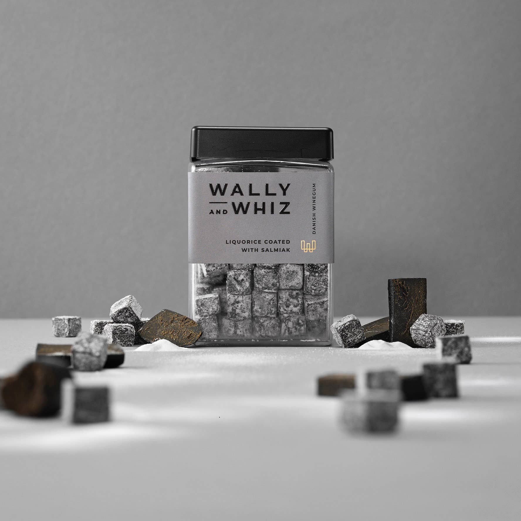 A container of Candy: Wally & Whiz Licorice With Salmiak 240g is surrounded by loose cubes on a surface. The label emphasizes the complementary flavors of licorice coated with salmiak. The monochromatic gray background perfectly showcases these salty licorice delights.