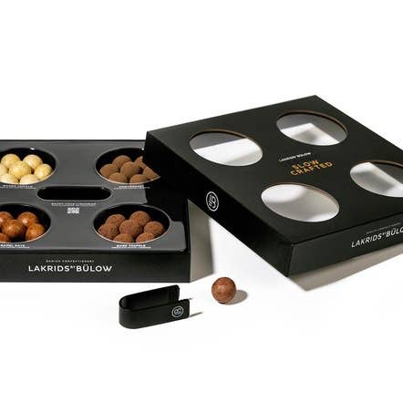 An open black box of Candy: Lakrids by Bülow - Organic Slow Crafted (120g) featuring assorted chocolate and licorice balls. A small black tong is placed beside one ball, inviting you to sample these responsibly crafted treats.