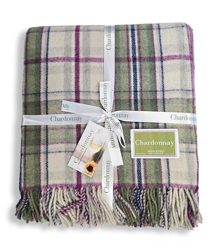 A folded Chardonnay Deluxe Tartan Blanket Merino Wool with green, purple, and cream checks, tied with a white ribbon labeled Chardonnay and a tag showing a wine glass and product details.