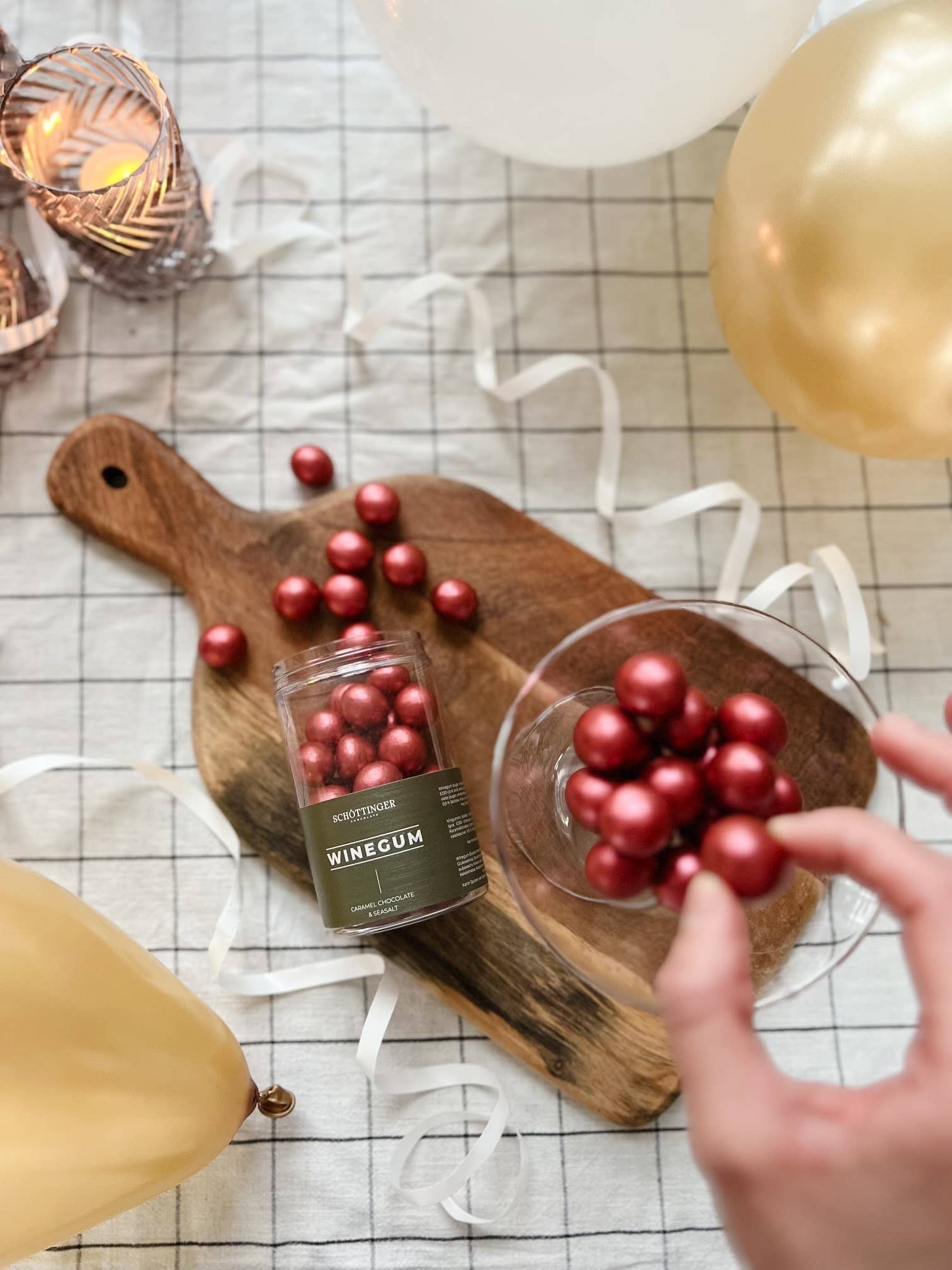 The Classic Winegum Seasalt & Caramel Chocolate candies in a jar and bowl on a wooden board with balloons, candles, and ribbons on a checkered surface shine like red amber under the light.