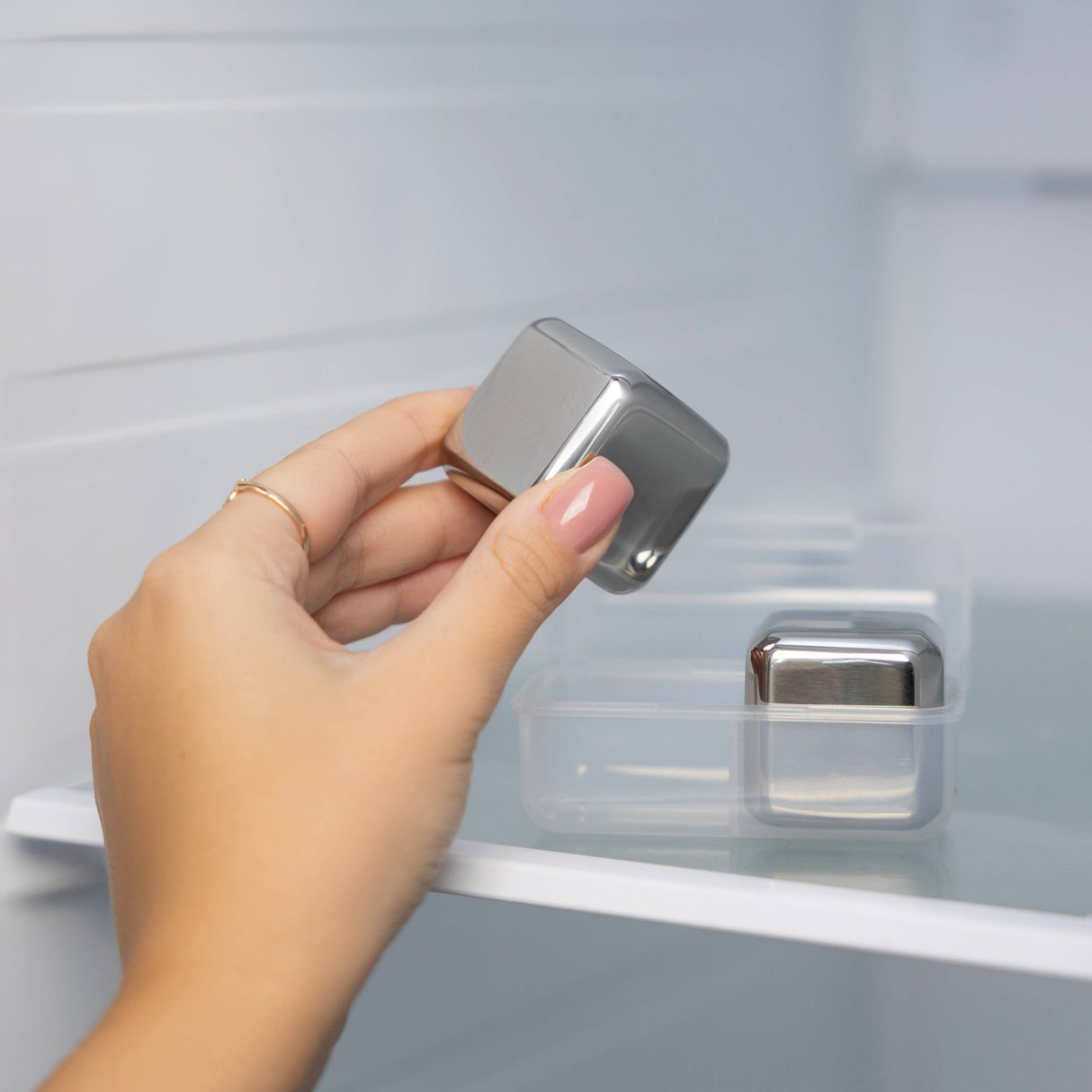 A hand places a Cubes: Stainless Steel Ice Cube (1.57" Square) from the Set of 2 into its plastic case and puts it in the refrigerator.