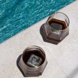 Two glasses with dark drinks and large Cubes: Stainless Steel Ice Cubes (Set of 2, 1.57" square) in a plastic case rest on a white poolside surface near blue water.
